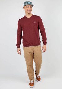 Pull à col en V bordeaux, pantalon khaki et baskets marron. Le pull a une texture subtile ; la casquette de baseball présente un motif géométrique.