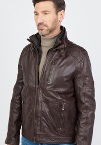 Bruine leren jas met een hoge kraag, ritsluiting aan de voorkant, meerdere zakken, schouderaccenten en een gladde textuur. Draagt over een beige coltrui.