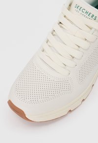 Valge sneaker perforeritud nahast ülemise, lamepeente ja pruuni kummist välistallaga. Bränding keelega rohelises värvitoonis.