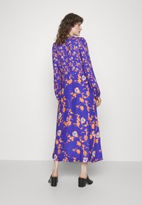 Vestido de manga longa, roxo, com padrões florais laranja e azul, um ajuste solto e comprimento até a meia panturrilha. Tecido suave. Saltos bloco pretos.