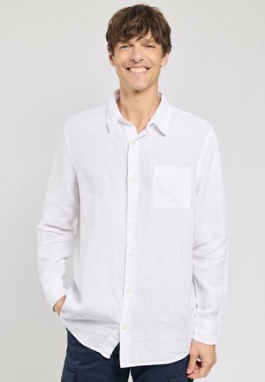Homme souriant aux cheveux bruns ébouriffés portant une chemise blanche à boutons avec une poche avant et un pantalon foncé, les mains dans les poches, fond gris clair.