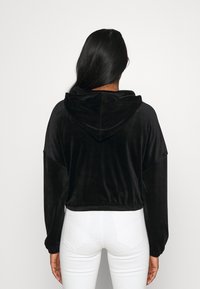 Sudadera negra corta de material suave y aterciopelado, con capucha ajustable y mangas sueltas, combinada con jeans blancos ajustados.