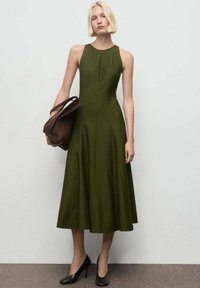 Vestido sin mangas en tejido de color verde oscuro con una textura suave. Presenta un escote redondeado y cae por debajo de la rodilla. Combinado con un bolso marrón.