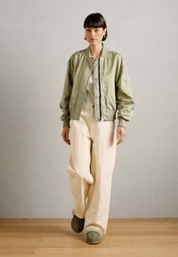 Licht olijfgroene bomberjack met geribbelde manchetten en kraag, gecombineerd met losse beige broek en grijs-groene slippers met een bontvoering.