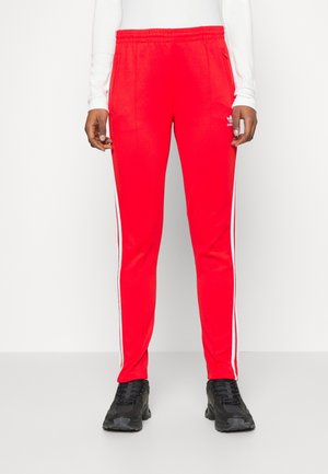 Pantalones deportivos rojos de Adidas con franjas laterales blancas, bolsillos con cremallera, combinados con zapatillas negras y una camiseta blanca de manga larga, mostrados de la cintura a los pies.