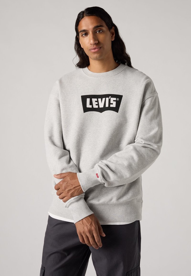Felpa grigia in pile con il logo nero di Levi's sul davanti. Presenta polsini e orlo a costine, e una piccola etichetta rossa di Levi's sul polsino.