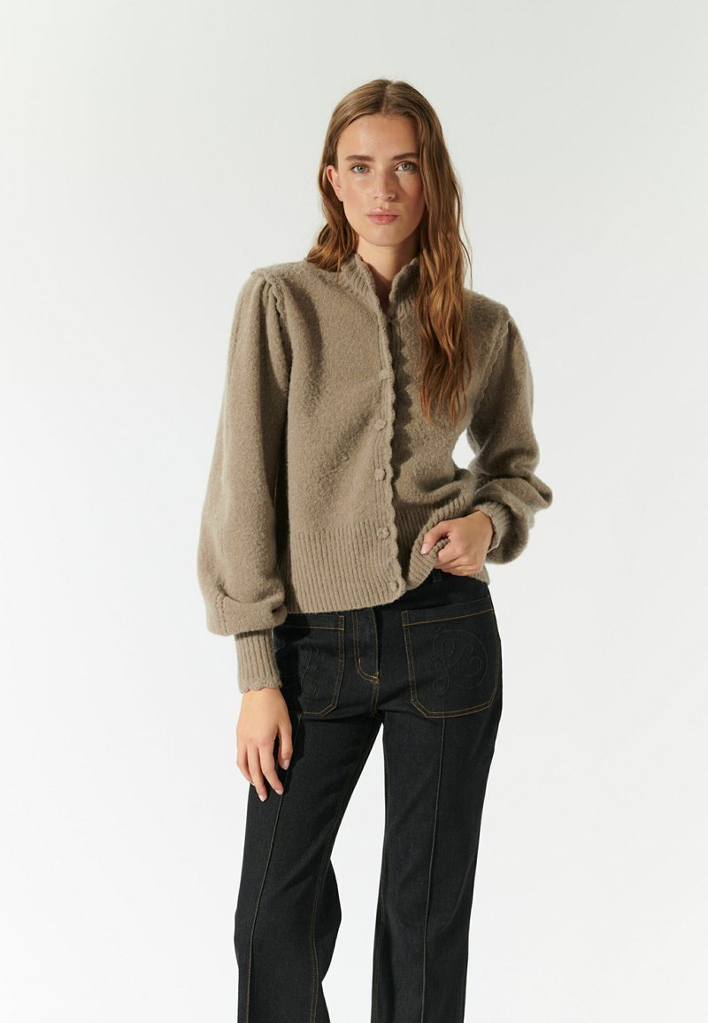 Lichtbruine gebreide cardigan met een gestructureerde afwerking, ronde kraag en knoopsluiting. Gepaard met donkerdenim broek met decoratieve stiksels.