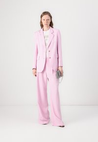 Traje a medida rosa con solapa en forma de muesca, doble botón y pantalones de pierna ancha. Combinado con una camisa con estampado y un pequeño bolso metálico.