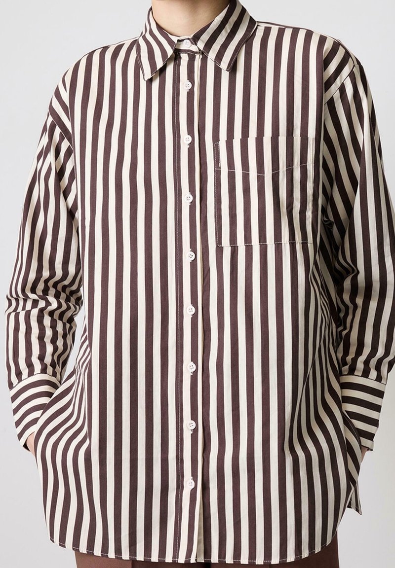 Personne portant une chemise à manches longues boutonnée avec des rayures verticales marron et blanches et une poche poitrine, les mains dans les poches latérales.