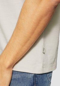 Close-up van de arm van een persoon naast de heup, gekleed in een wit shirt met het label "s.Oliver" en blauwe spijkerbroek.