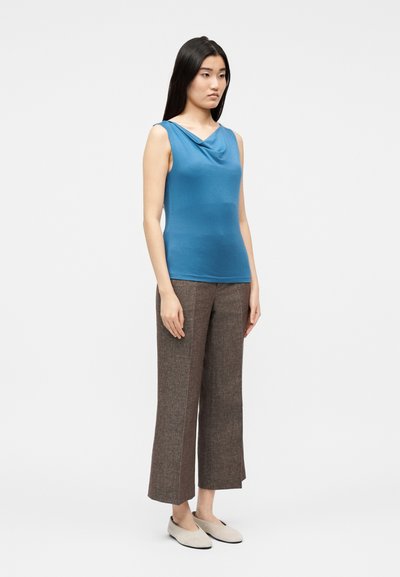 Γυναίκα όρθια, φορώντας μπλε αμάνικη μπλούζα με λαιμόκοψη draped, καφέ φαρδιά cropped παντελόνια και μπεζ επίπεδα παπούτσια, σε απλό φόντο.