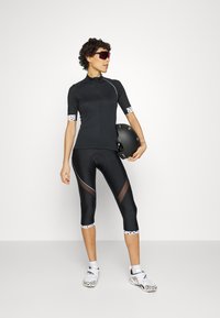 Camisola de ciclismo preta de mangas curtas, com punhos brancos com bolinhas; combinada com leggings capri pretas que apresentam painéis em rede. Os acessórios incluem óculos de sol e um capacete.