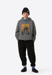 Hall hallipluus kirjaga "NEVER STOP EXPLORING" oranžis värvis, mustad dressipüksid ja must müts. Modell kannab siniseid sokke ja musti sussisid.