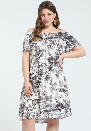 Vrouw draagt een witte off-shoulder jurk met zwarte botanische en dierenprint, poseert met één hand op de heup en lichtbruin golvend haar.