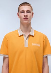 Orange polo skjorte i tekstureret stof med beige krave og knaplukning, der har et lille hvidt broderet logo på venstre bryst.