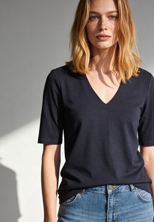 Junge Frau mit schulterlangen blonden Haaren, die ein schwarzes V-Ausschnitt-Kurzarmshirt und blaue Jeans trägt, steht vor einem einfarbigen Hintergrund.