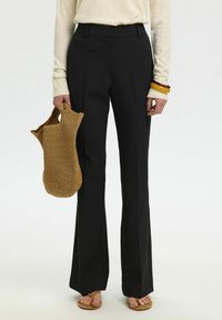 Femme portant un pantalon évasé noir taille haute, un haut à manches longues crème, des sandales marron, tenant un sac tissé marron, avec des bracelets au poignet droit.