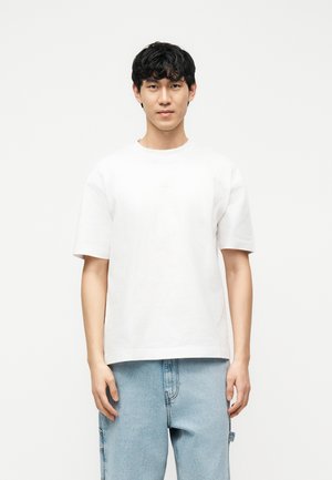 Noor mees lühikeste mustade juustega, kandes tavalist valget T-särki ja helehalli teksapükse, seismas valge tausta ees.