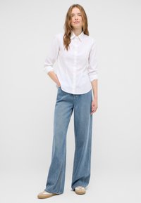 White button-up shirt with a collar, long sleeves rolled up, paired with wide-leg light blue jeans and beige slip-on shoes.