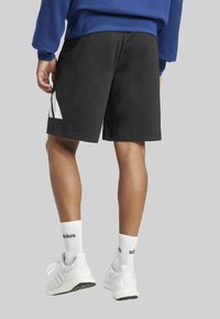 Black athletic shorts with white triangular design accents, paired with white sneakers and ribbed white socks. Background is plain gray.