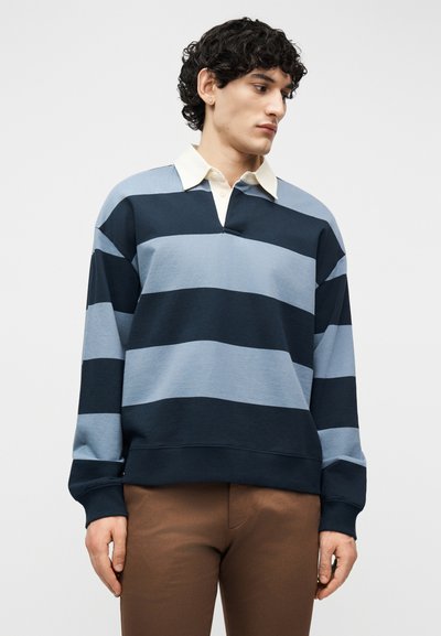 Άνδρας που φοράει μπλε και σκούρο μπλε ριγέ ράγκμπι πουκάμισο με λευκό γιακά και καφέ παντελόνι, κοιτάζει προς τα δεξιά του.