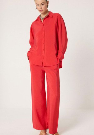 Kobieta stojąca z rękami w kieszeniach, ubrana w luźną czerwoną koszulę na guziki i pasujące szerokie spodnie oraz brązowe sandały.