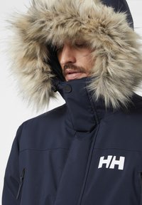 Námorníková zimní bunda s kapucí a umělou kožešinou, s vysokým límcem, zipovými kapsami a bílým logem na hrudi.