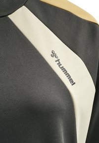 Gros plan sur un tissu de vêtements de sport en blocs de couleur gris foncé, beige et crème avec un petit logo "hummel" et une icône d'abeille.