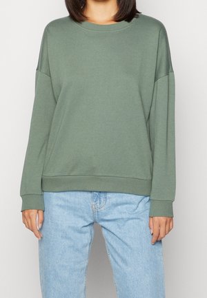 Vrouw draagt een effen olijfgroene trui met lange mouwen en lichtblauwe jeans met hoge taille, staand tegen een witte achtergrond.