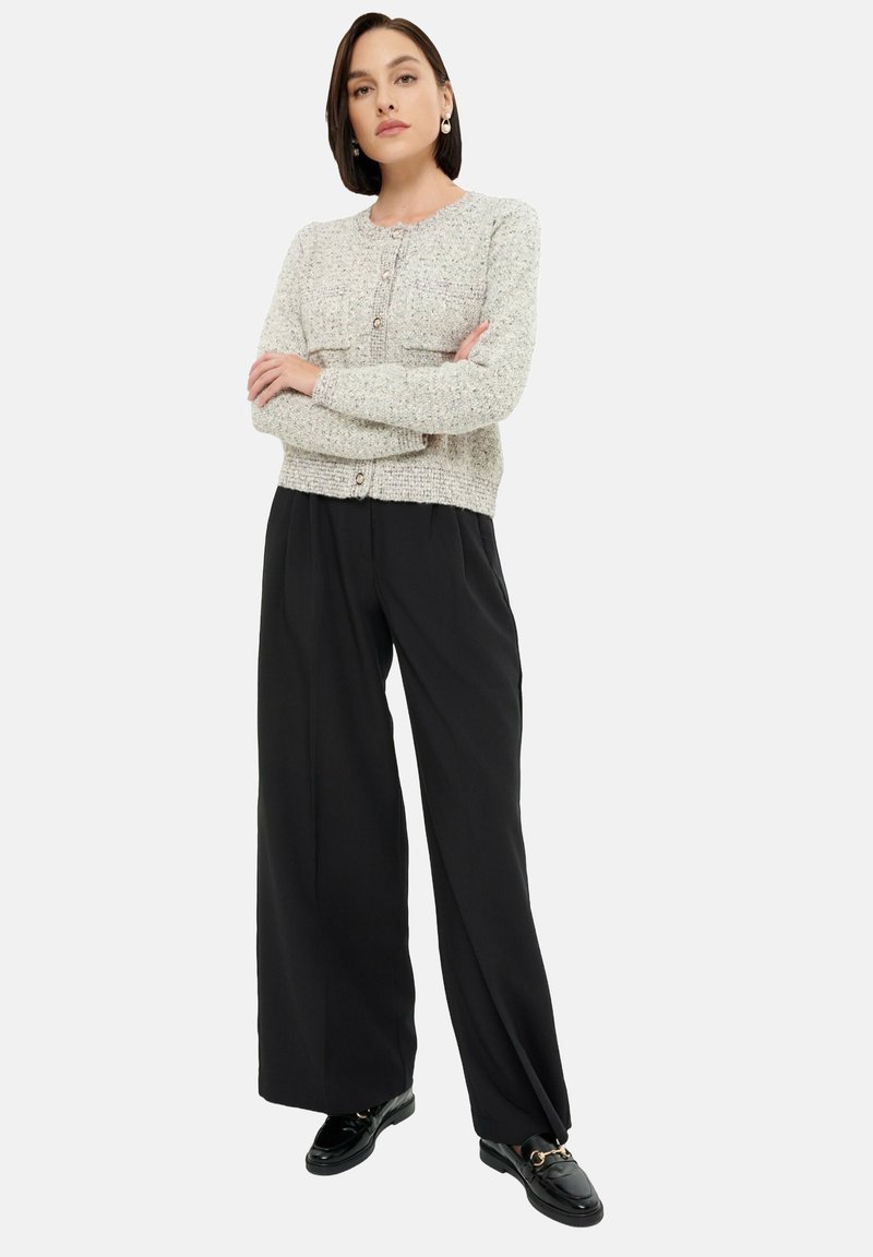 Jasnoszary sweter dzianinowy z dwoma kieszeniami, zapinany na guziki z przodu, w zestawieniu z szerokimi czarnymi spodniami i czarnymi mokasynami. Gładka faktura.