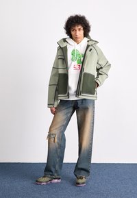 Hellgrüne wasserdichte Jacke mit schwarzen Akzenten, mehreren Taschen, über einem weißen Hoodie mit grünem Blattmuster getragen, kombiniert mit ausgefransten Jeans und olivfarbenen Sneakers.