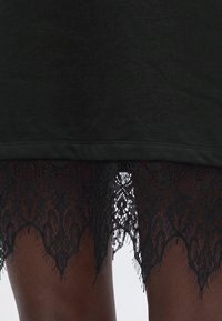 Robe en tissu noir avec ourlet en dentelle noire festonnée sur peau foncée, montrant un motif floral en dentelle détaillé et une légère transparence.