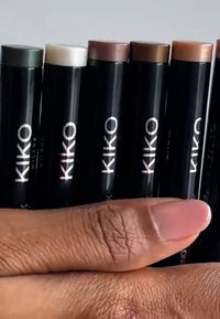 Zestaw ośmiu sztyftów do makijażu KIKO Milano w czarnych tubkach z srebrnymi logo. Kolory obejmują zielony, biały oraz różne odcienie brązu.