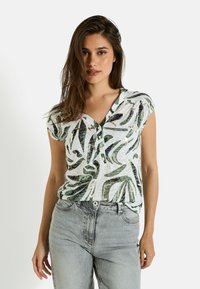 Blusa de manga corta, ligera, en blanco con patrones de hojas verdes, con escote en V y detalles de botones, acompañada de jeans de mezclilla gris.