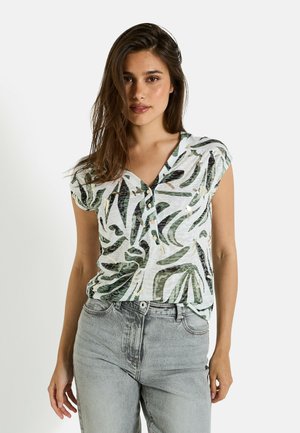 Blouse légère à manches courtes de couleur blanche avec des motifs de feuilles vertes, présentant un col en V et des détails de boutons, associée à un jean en denim gris.