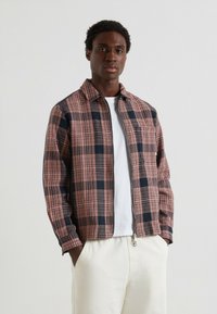 Kurtka w kratę w odcieniach różowego i granatowego, z zapięciem na zamek i kołnierzowym dekoltem, w zestawieniu z białą koszulą i jasnymi spodniami.