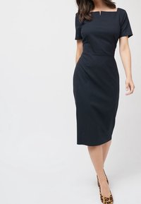 Marineblaue enganliegende Kleid mit kurzen Ärmeln, quadratischem Ausschnitt und Nahtdetails. Bleistiftsilhouette reicht bis knapp unter die Knie.