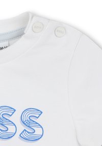 T-shirt bianco in cotone con scritta blu a righe "BOSS." Presenta due bottoni a pressione bianchi sulla spalla sinistra per la chiusura. Texture liscia.