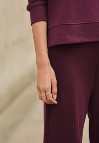 Bluza burgundă cu un finisaj texturat, asortată cu pantaloni în aceeași nuanță, având o siluetă netedă și o mână cu inele argintii.