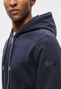Tamno plavi zip-up hoodie napravljen od mekanog materijala. Ima veliki kapuljaču, rebraste manžetne i oznaku brenda na lijevoj ruci. Glatka tekstura.