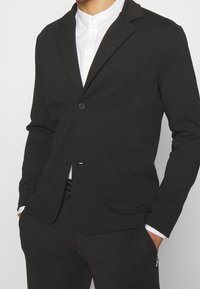 Sort blazer i blødt stof med et streamlined design, to-knaps lukning og forlommer. Båret med en hvid skjorte nedenunder.