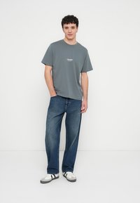 Grijs katoenen T-shirt met ronde hals, voorzien van een klein wit logo. Gecombineerd met losse donkerblauwe jeans en witte sneakers met zwarte accenten.