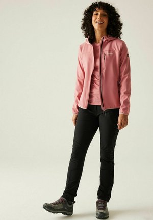 Frau mit lockigem Haar, die eine rosa Reißverschlussjacke, passendes Shirt, schwarze Hose und graue Wanderschuhe trägt, stehend und lächelnd.