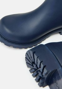 Námornícky modré gumové čižmy so hladkým povrchom, hrubou textúrovanou podrážkou a hlbokými drážkami pre trakciu. Na vrchu mierne zakrivený tvar.