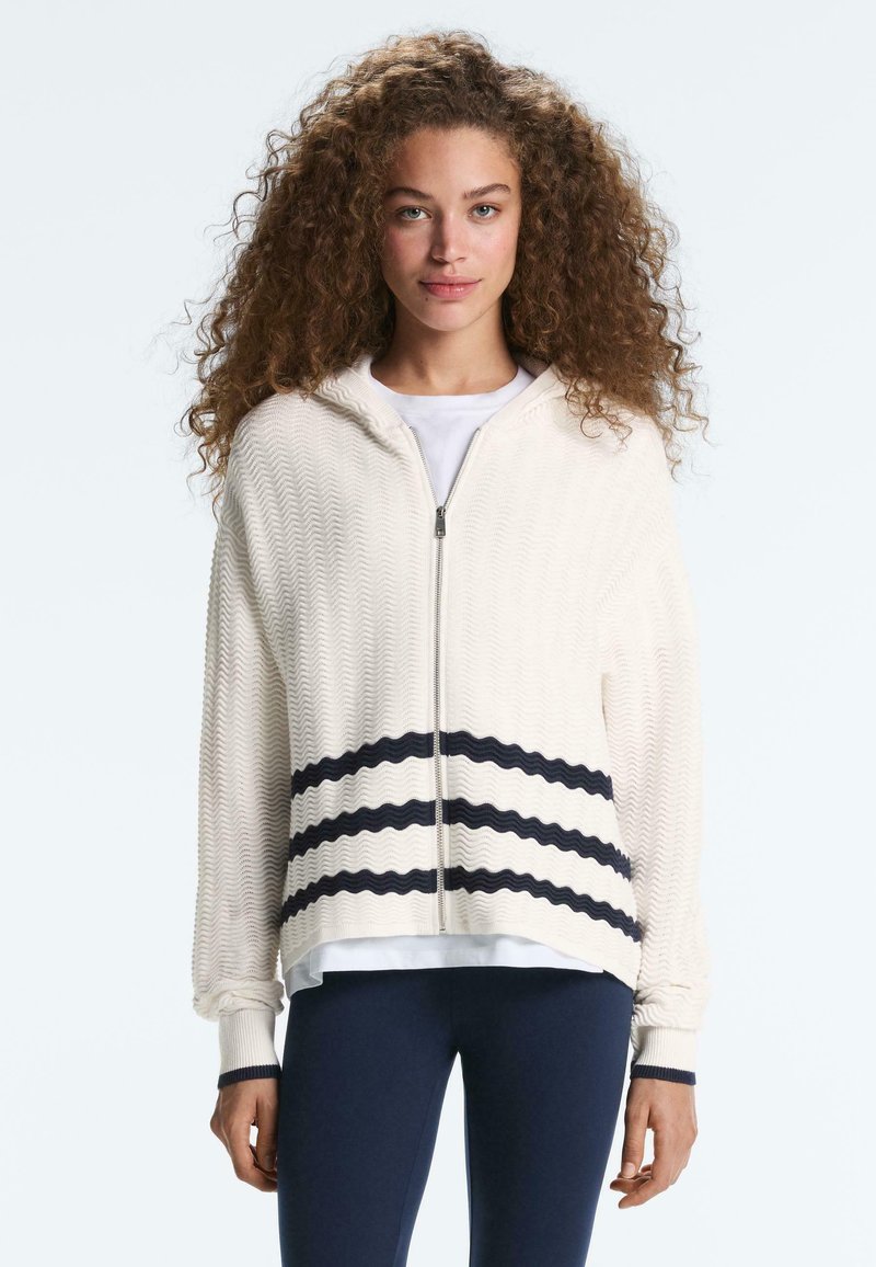 Femme aux cheveux bouclés portant un sweat à capuche blanc texturé zippé avec des rayures ondulées noires et un pantalon bleu marine, debout sur un fond uni.