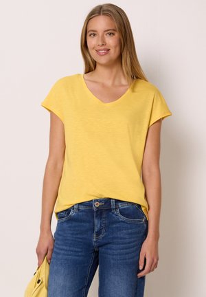 Smilende kvinde med langt blond hår iført en gul kortærmet skjorte og blå jeans, der holder en sammenfoldet gul jakke i den ene hånd.