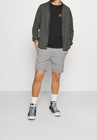 Camicia grigia a maniche lunghe sopra una maglietta nera, abbinata a pantaloni corti grigi chiari, calze bianche e sneaker alte nere. Design semplice con un look casual.