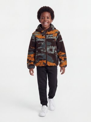 Enfant souriant marchant, portant une veste polaire zippée à capuche avec un motif camouflage orange, marron et vert, pantalon noir et baskets blanches.