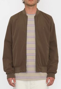 Veste aviateur marron à finition lisse, fermeture éclair, poignets côtelés et ourlet ajusté, superposée sur une chemise légère rayée.