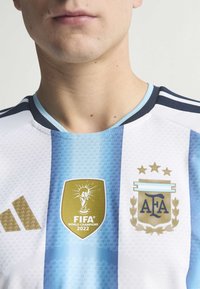 Bijela i svijetloplava prugasta nogometna majica s zlatnim logom FIFA Svjetski prvak 2022, AFA amblemom i crnim Adidas brandingom.
